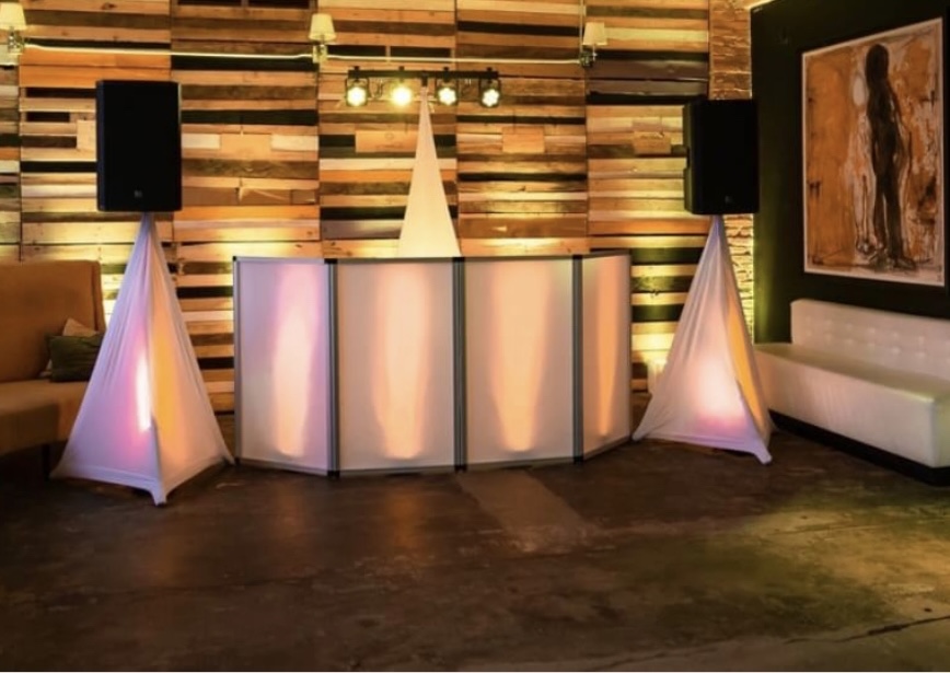 White DJ facade and covered speaker stands with warm uplighting on a wood wall